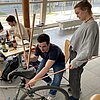 Das Bild zeigt einen Studenten, der an einem Fahrrad sitzt und einen Gepäckträger aus Holz baut. Neben ihm steht eine Studentin.