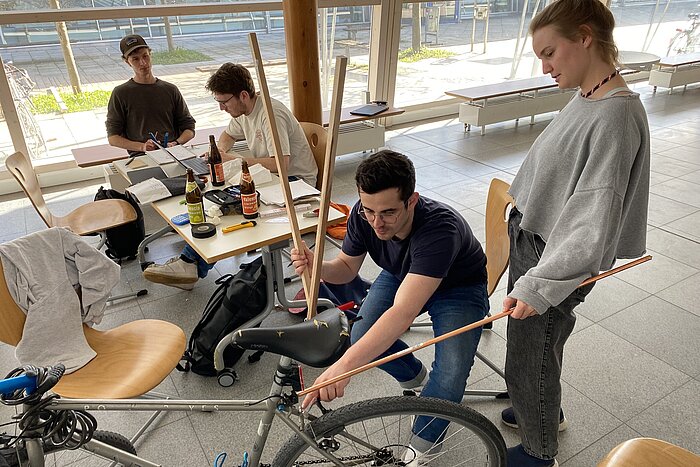 Das Bild zeigt einen Studenten, der an einem Fahrrad sitzt und einen Gepäckträger aus Holz baut. Neben ihm steht eine Studentin.