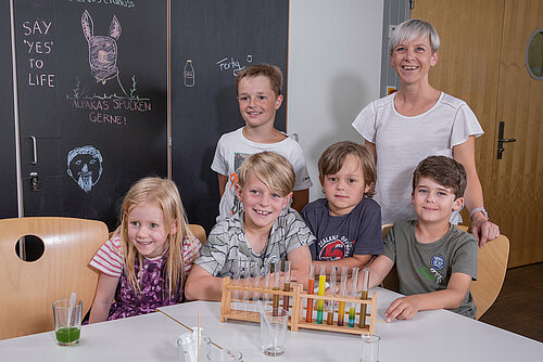 Eine Frau betreut 5 lachende Kinder im Klassenzimmer. Sie haben Spielzeug auf dem Tisch stehen und eine Tafel zum Zecichnen und Schreiben.
