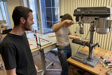 Das Bild zeigt eine Studentin, die an einer Maschine ein Stück Holz bearbeitet. Neben ihr steht ein Student. 