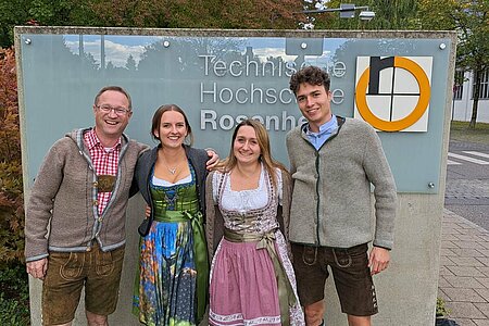 Two men in leather pants and two women in traditional Bavarian dress: Dirndl in front of the TH Rosenheim Logo