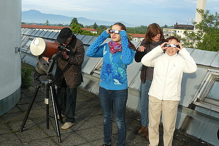 Kinder beobachten den Venusdurchgang mit speziellen Brillen und ein Mann beobachtet ihn durch ein Teleskop.