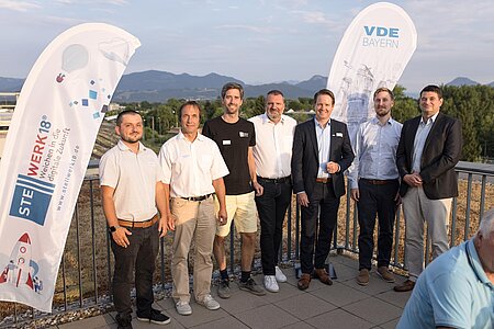 sieben männliche Teilnehmer der Veranstaltung stehen nebeneinander für ein Gruppenfoto auf dem Dach des Gebäudes am Geländer.