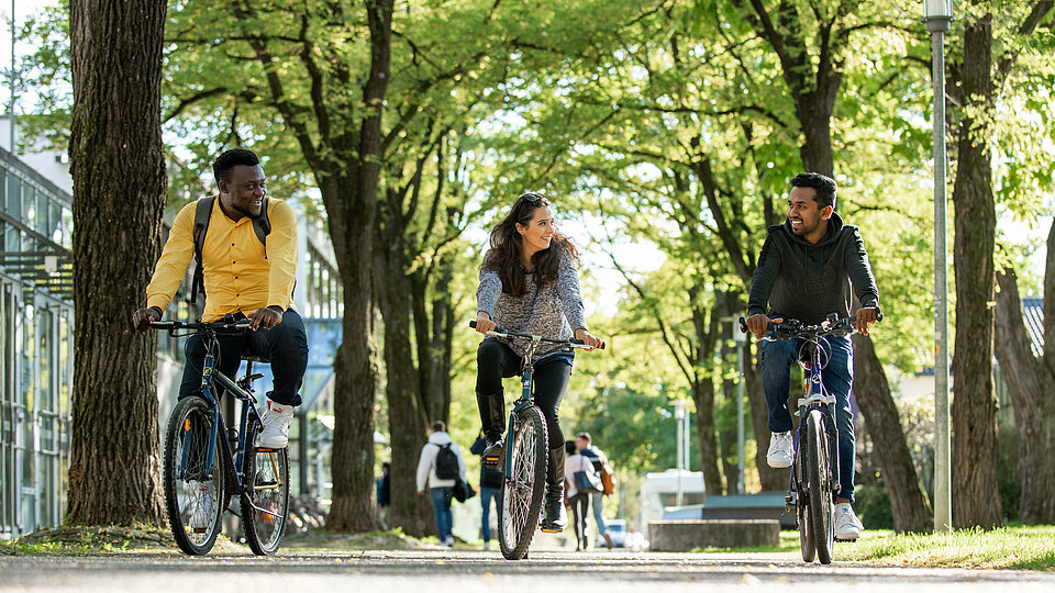 [Translate to English:] Drei Studierende fahren mit dem Rad durch den Campus.