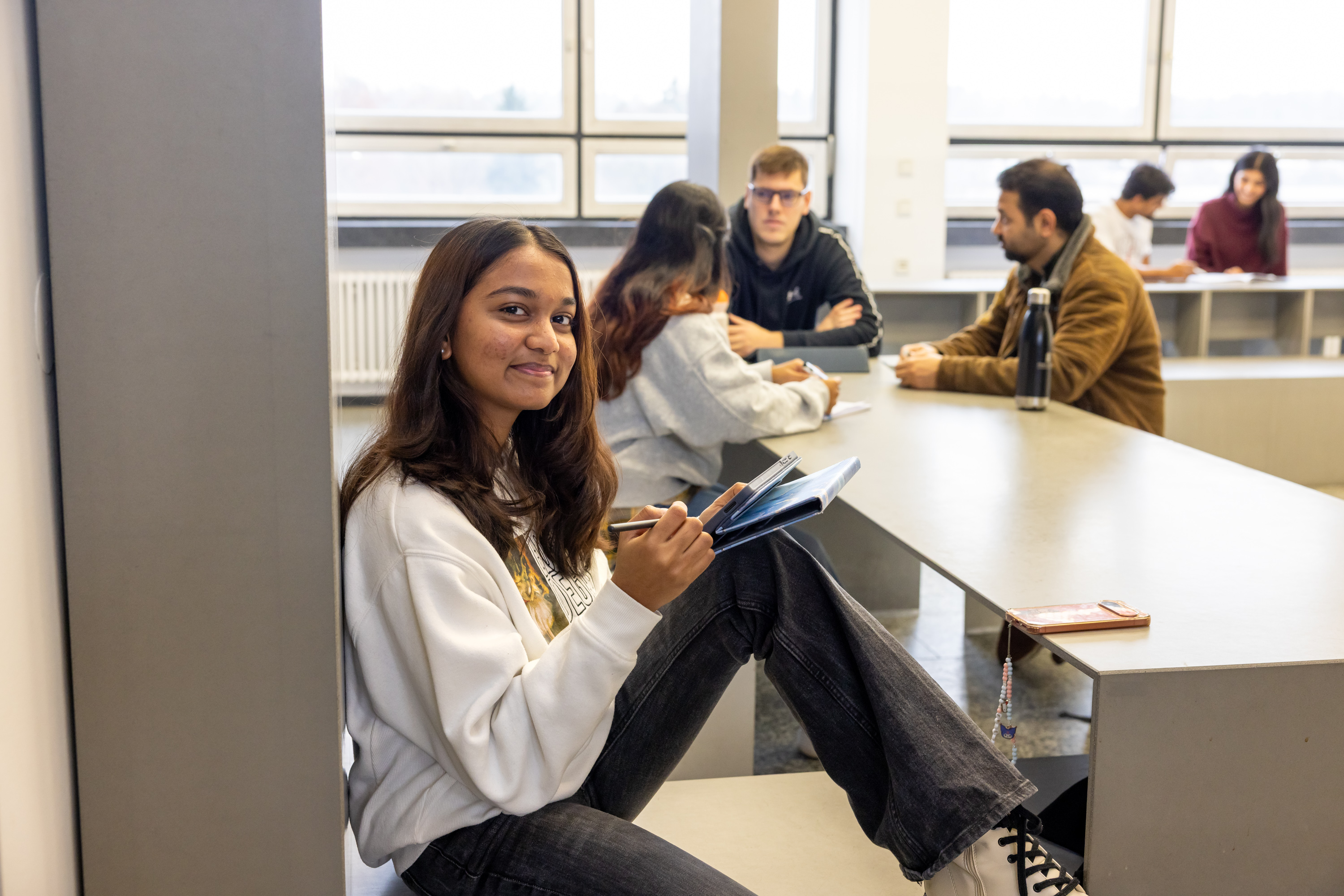 Studentin im Vordergrund sitzt an einer Säule lehnend. Im Hintergrund ist eine Studierendengruppe beim Austausch zu sehen.