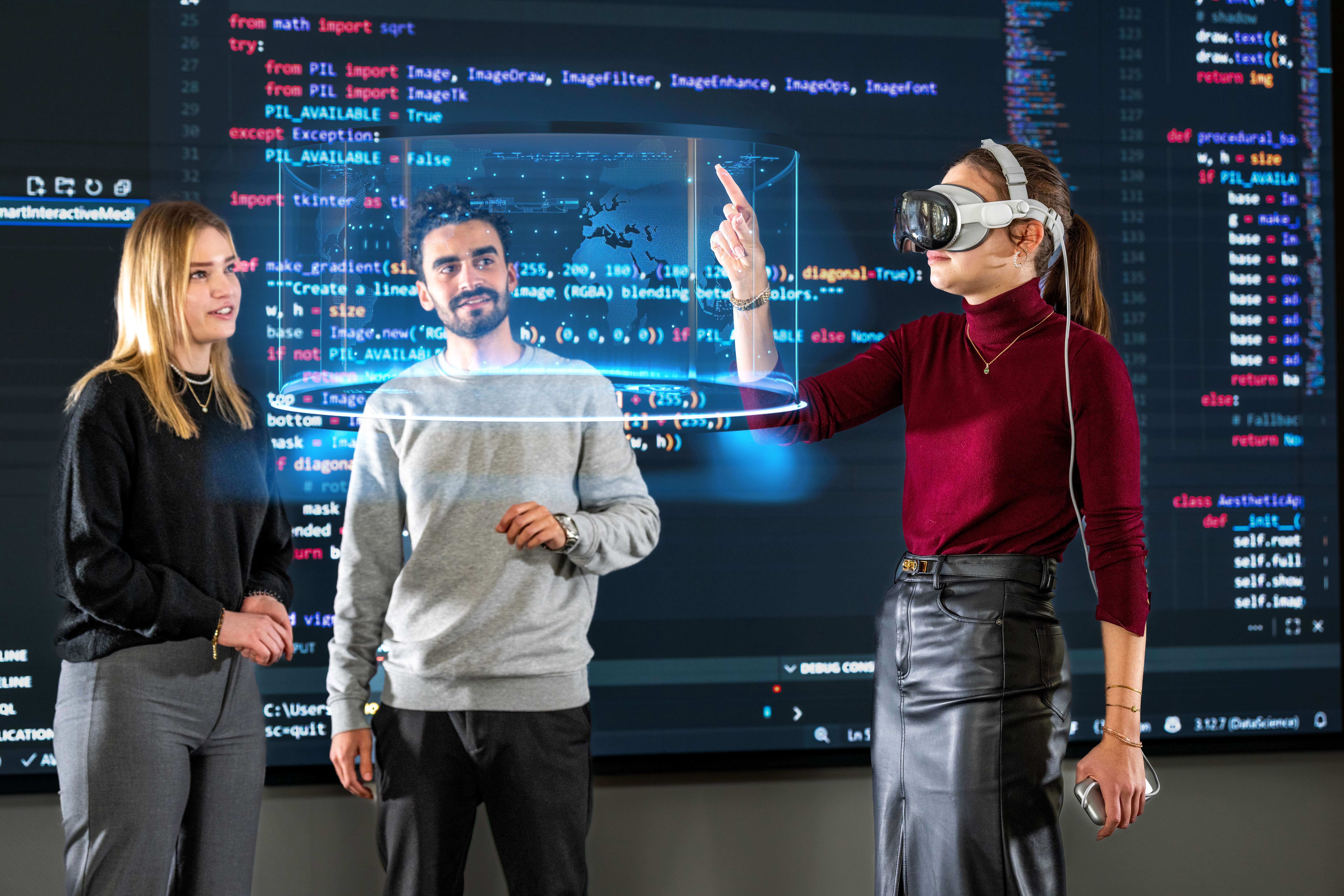 Studentin mit einer VR-Brille zeigt auf ein Hologramm. Eine weitere Studentin und ein Student schauen auf das Hologramm.
